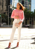 Woman in a pink textured jacket and white pants standing in an urban setting with modern buildings.