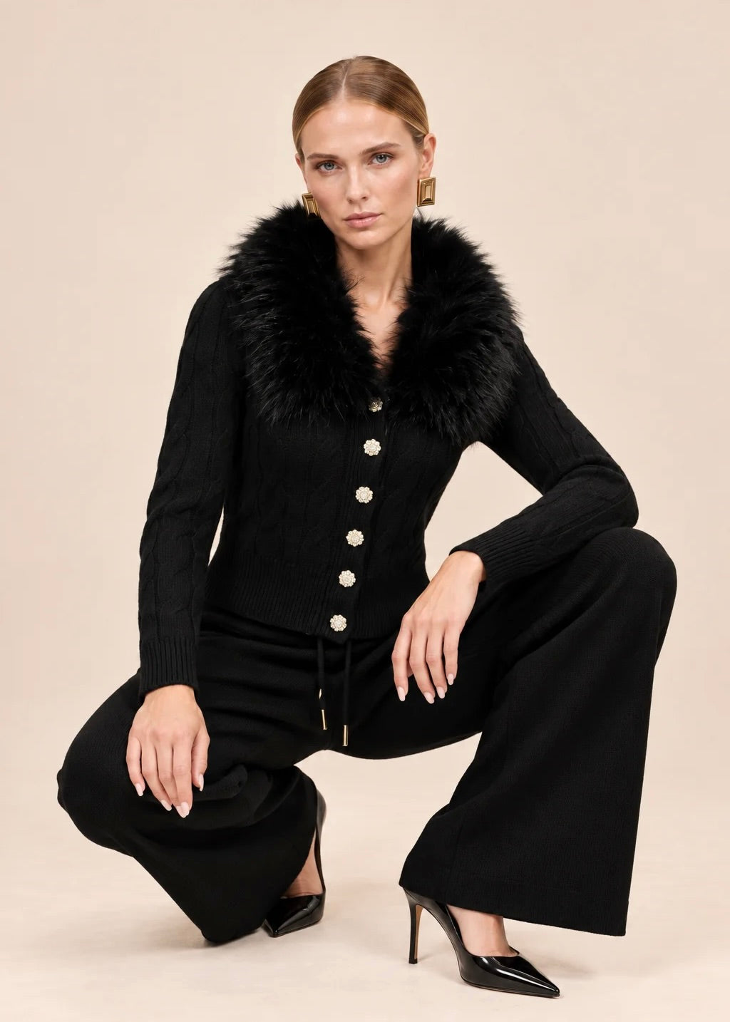 Woman crouching in black knit cardigan and wide-leg pants set with faux fur collar